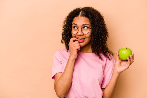 Młoda Afroamerykanka trzymająca jabłko odizolowane na beżowym tle rozluźnia się myśląc o czymś patrząc na przestrzeń kopii. - Zdjęcie, obraz
