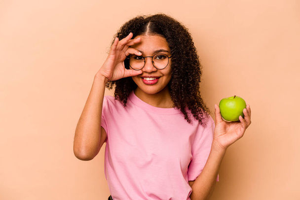 Młoda Afroamerykanka trzymająca jabłko odizolowane na beżowym tle podekscytowana trzymaniem ok gestu na oku. - Zdjęcie, obraz