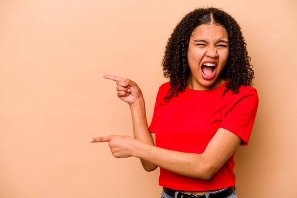 Junge afrikanisch-amerikanische Frau isoliert auf beigem Hintergrund zeigt mit den Zeigefingern auf einen Kopierraum und drückt damit Begeisterung und Sehnsucht aus. - Foto, Bild