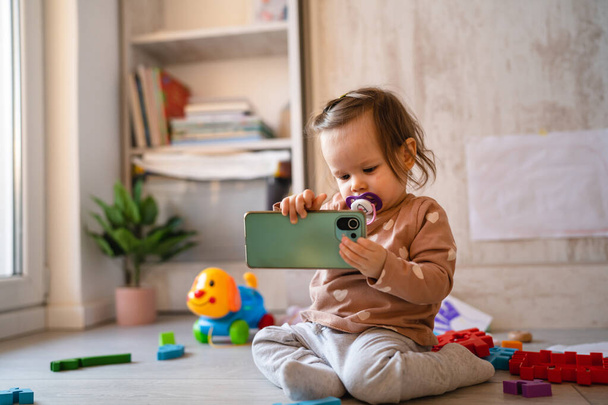 One baby small caucasian infant girl playing on the floor at home copy space holding mobile phone smartphone using to call or watch video in day front view - Фото, изображение
