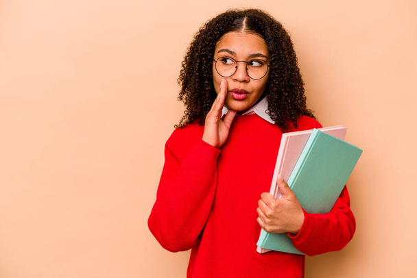 Giovane studentessa afroamericana isolato su sfondo beige sta dicendo un segreto caldo frenante notizie e guardando da parte - Foto, immagini