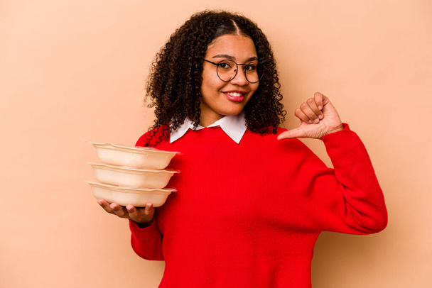 Joven mujer afroamericana sosteniendo tupperware aislado sobre fondo beige se siente orgulloso y seguro de sí mismo, ejemplo a seguir. - Foto, Imagen