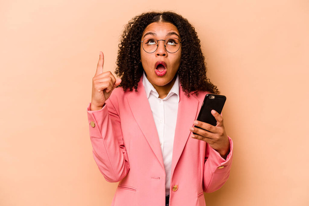 Young African American woman holding mobile phone isolated on beige background pointing upside with opened mouth. - Foto, Bild