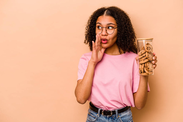Joven mujer afroamericana sosteniendo un tarro de galletas aislado sobre fondo beige está diciendo una noticia secreta de frenado caliente y mirando a un lado - Foto, Imagen