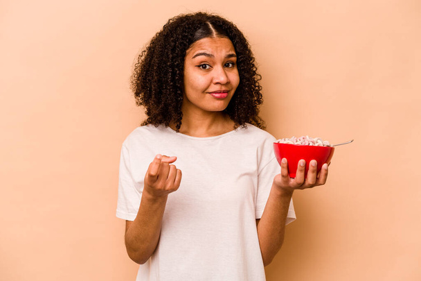 Young African American woman holding a bowl of cereals isolated on beige background pointing with finger at you as if inviting come closer. - Foto, afbeelding