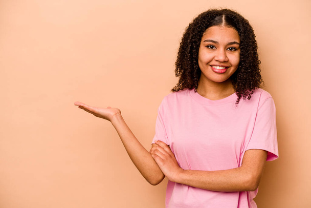 Jeune femme afro-américaine isolée sur fond beige montrant un espace de copie sur une paume et tenant une autre main à la taille. - Photo, image