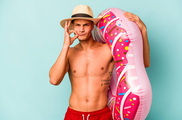 Joven hombre caucásico sosteniendo un donut inflable aislado sobre fondo azul con los dedos en los labios manteniendo un secreto. - Foto, Imagen