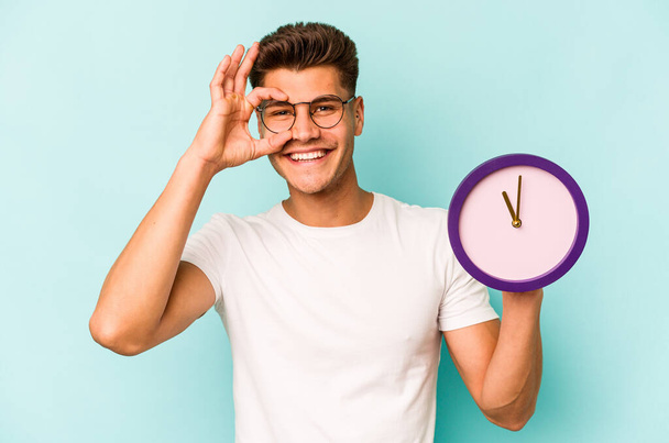 Young caucasian man holding a clock isolated on blue background excited keeping ok gesture on eye. - Fotó, kép