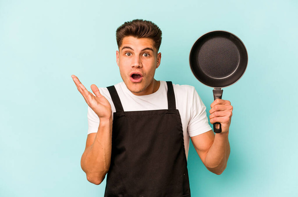 Young caucasian cooker holding frying pan isolated on blue background surprised and shocked. - Photo, Image