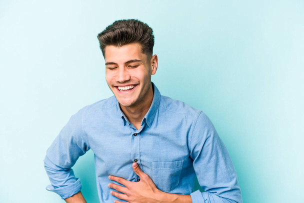 Young caucasian man isolated on blue background laughing keeping hands on heart, concept of happiness. - 写真・画像