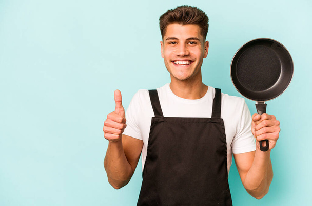 Young caucasian cooker holding frying pan isolated on blue background smiling and raising thumb up - Фото, изображение