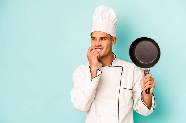 Young caucasian chef man holding flying pan isolated on blue background relaxed thinking about something looking at a copy space. - Photo, Image