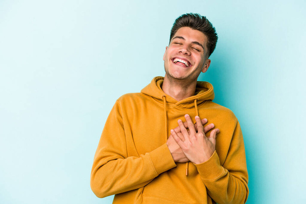 Молодий кавказький чоловік ізольований на синьому фоні сміється тримаючи руки на серці, концепція щастя
. - Фото, зображення
