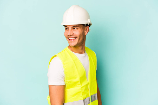 Young laborer caucasian man isolated on blue background looks aside smiling, cheerful and pleasant. - Foto, Imagem