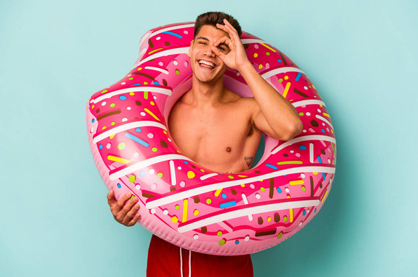Young caucasian man holding an inflatable donut isolated on blue background excited keeping ok gesture on eye. - Fotó, kép