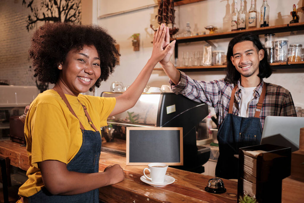 Two cafe business startup partners and friends, African American female, and Thai male baristas look at camera, hand in hand, and cheerful smiles at coffee shop counter bar, happy service staff job. - Photo, Image