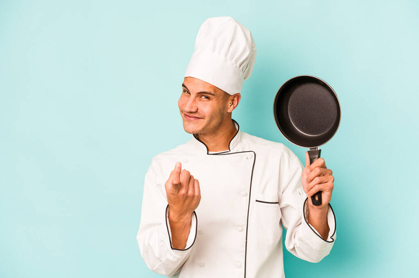Молодой кавказский шеф-повар держит летающую сковороду на голубом фоне, указывая пальцем на тебя, словно приглашая подойти ближе.. - Фото, изображение