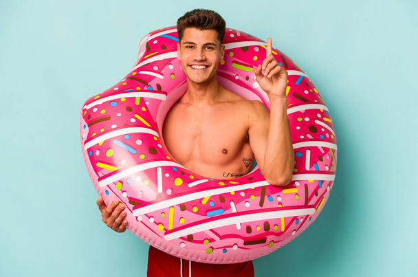 Young caucasian man holding an inflatable donut isolated on blue background showing number one with finger. - Foto, afbeelding