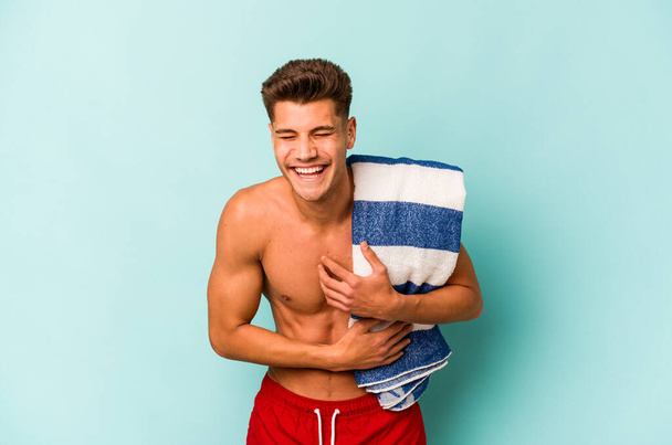 Young caucasian man holding beach towel isolated on blue background laughing and having fun. - Foto, Imagem