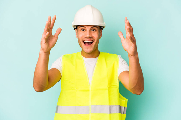 Young laborer caucasian man isolated on blue background receiving a pleasant surprise, excited and raising hands. - 写真・画像