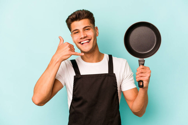 Young caucasian cooker holding frying pan isolated on blue background showing a mobile phone call gesture with fingers. - Фото, изображение