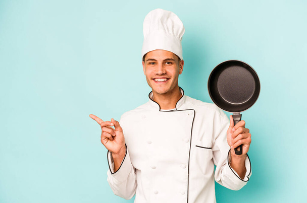 Young caucasian chef man holding flying pan isolated on blue background smiling and pointing aside, showing something at blank space. - Foto, imagen