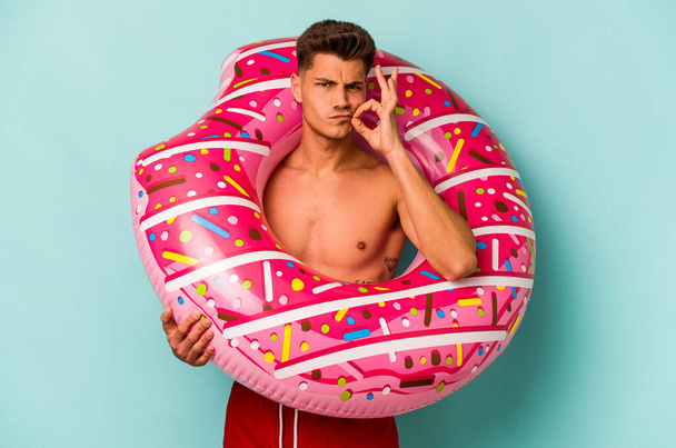 Young caucasian man holding an inflatable donut isolated on blue background with fingers on lips keeping a secret. - Фото, зображення