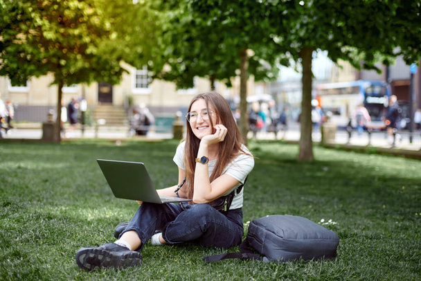 Uma mulher senta-se no relvado com um portátil a sonhar. Uma mulher trabalha remotamente com um laptop ao ar livre no parque - Foto, Imagem