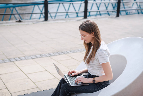 Una joven trabaja en su portátil, sentada en un banco moderno en el paseo marítimo de Newcastle - Foto, imagen