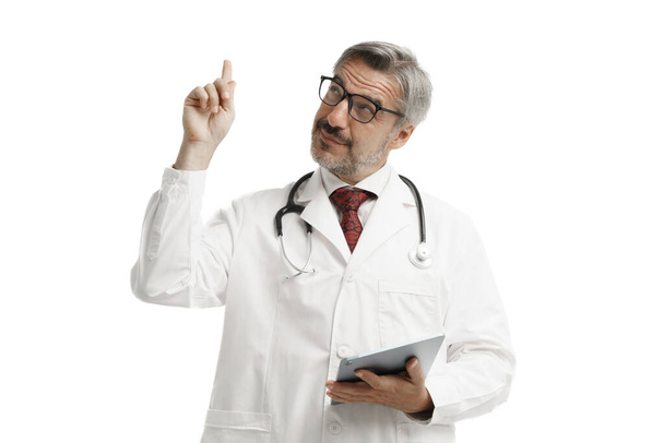 Portré magabiztos fehér férfi orvos köpenyben egyenruha sztetoszkóppal és tabletta mutat a kezét, és felnéz a tetejére, miközben állni fehér háttér, orvosi bemutató koncepció. - Fotó, kép