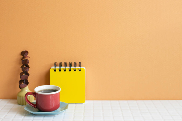 Filiżanka kawy i notatnik, suchy eukaliptus na białym stole. pomarańczowy tło ściany. przestrzeń kopiowania - Zdjęcie, obraz
