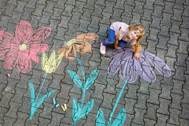Pequena pintura menina pré-escolar com giz colorido flores no chão no quintal. Criança feliz positiva desenhando e criando imagens no asfalto. Atividades criativas de crianças ao ar livre no verão. - Foto, Imagem