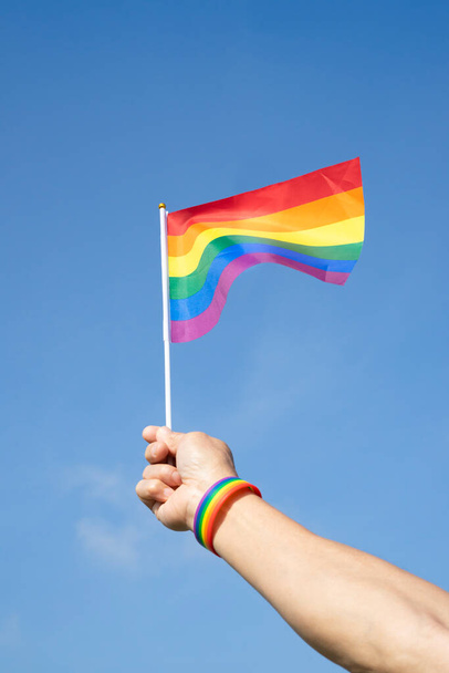 Мужская рука носит браслет ЛГБТ-радуги размахивает радужным флагом ЛГБТ на фоне голубого неба (избирательное внимание), концепция движения за равенство ЛГБТ-сообщества - Фото, изображение