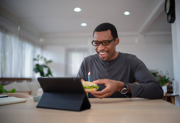 Happy handsome African American man is holding a hamburger celebrating birthday through video call virtual party with friends by a tablet distance at home. Modern Holiday Celebration - Photo, Image