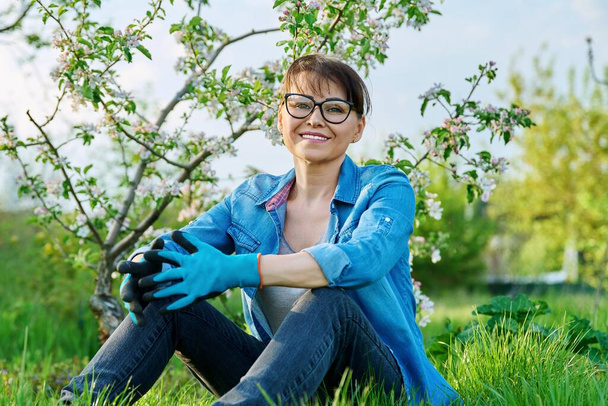 Positive beautiful mature female gardener sitting on the grass with blossoming apple tree. - Фото, зображення