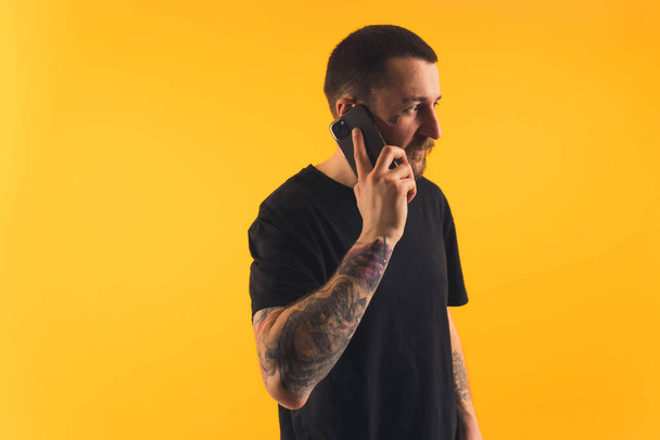 Young man talking on the phone, tattooed arms, looking away studio shot copy space isolated yellow background  - Foto, Imagen
