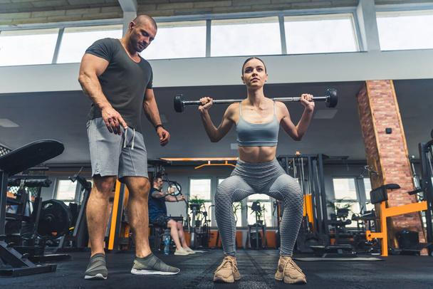 Front full body indoor shot of two caucasian people at gym - a male trainer and a sports girl - training weightlifting with small barbell. - Photo, Image