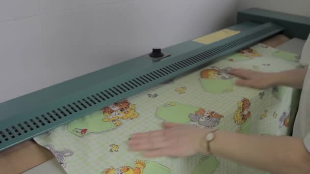 A háziasszony tiszta babaágyneműt tesz a nagy vasalógépbe. Női kezek, akik friss mosott ruhákat viselnek a mosodában. - Felvétel, videó