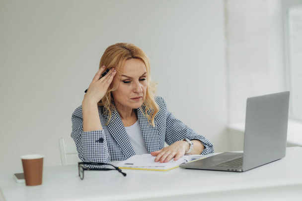 Tired senior business woman is using laptop for work indoor. A beautiful older lady has headache from overworking. Mature woman 50s - Фото, изображение