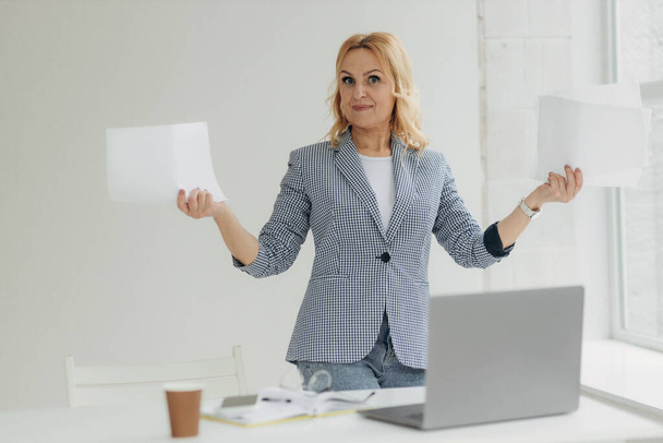 Skoncentrowana bizneswoman w średnim wieku stojąca w przytulnym biurze i analizująca papiery. Dojrzała kobieta po 50-tce - Zdjęcie, obraz