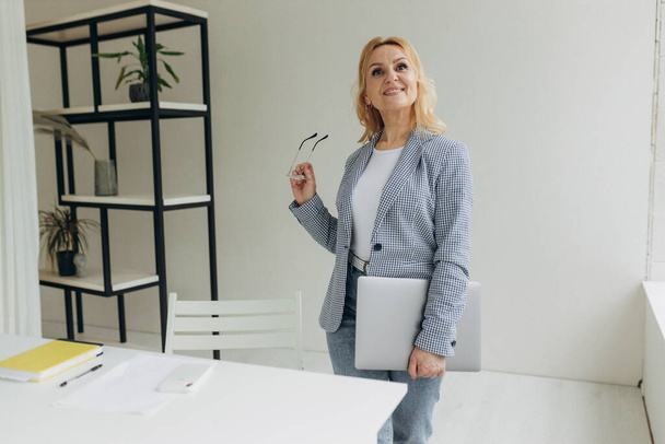 Joyful blonde-hair business woman in eyeglasses is looking at the away, holding laptop in arms and smiling. Beautiful middle aged lady in stylish wear stands in office. Mature woman 50s - Foto, Imagem
