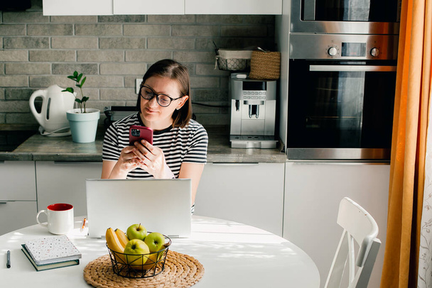 Freelancer woman with dark short hair wearing glasses using phone and laptop while working from home on kitchen. - Foto, Imagem