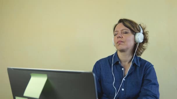 Une femme de quarante ans vêtue d'une chemise bleue écoute de la musique dans de grands écouteurs filaires blancs avec des ordinateurs portables à la table. Portrait au ralenti d'une femme adulte gaie - Séquence, vidéo