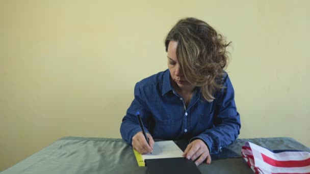 Une femme de quarante ans en chemise en denim écrit avec un stylo dans un carnet de notes en papier alors qu'elle est assise à une table. Portrait d'une femme adulte - Séquence, vidéo