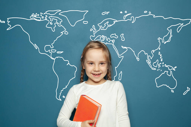 Heureux écolier enfant avec livre debout sur fond bleu avec carte du monde - Photo, image
