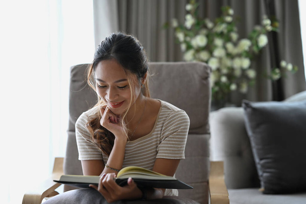 Glückliche Millennial-Frau liest Buch, während sie auf beigem Sessel in der Wohnung sitzt. - Foto, Bild