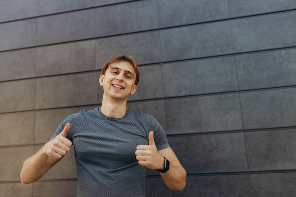 Photo of a strong athlete standing isolated over gray wall background. Looking at camera, pointing thumb up. excited cheerful guy. - Fotó, kép