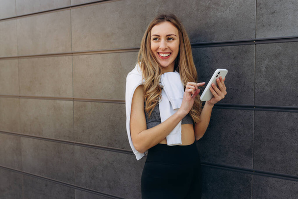 Beautiful woman wearing gray sporty top while holding smart phone in hands, having light hair, using cell phone while posing outdoor near gray wall. Copy space - Photo, Image