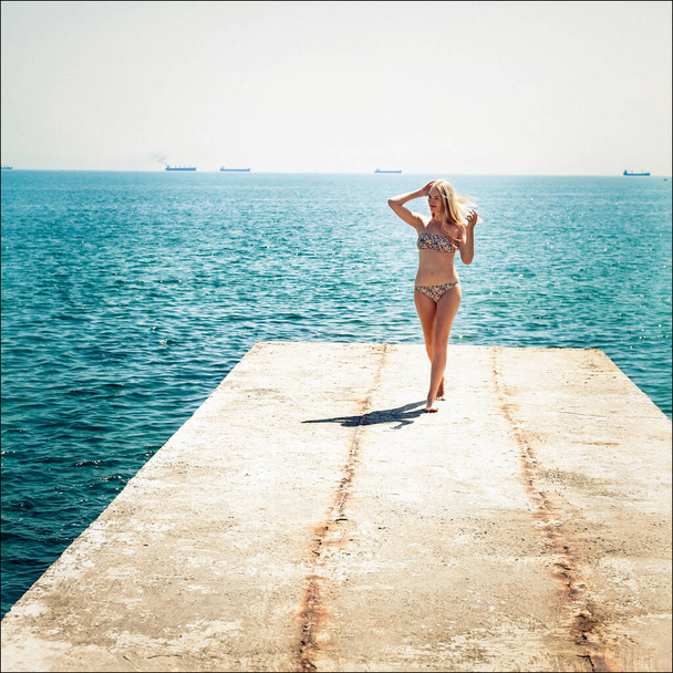 Стройная девушка с длинными светлыми волосами в стильном купальнике идет вдоль пирса у моря, в ветреный солнечный день. Корабли видны вдалеке. - Фото, изображение