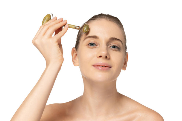 Portrait of young beautiful woman taking care after skin, massaging forehead with cosmetic roller tool isolated over white studio background - Photo, image